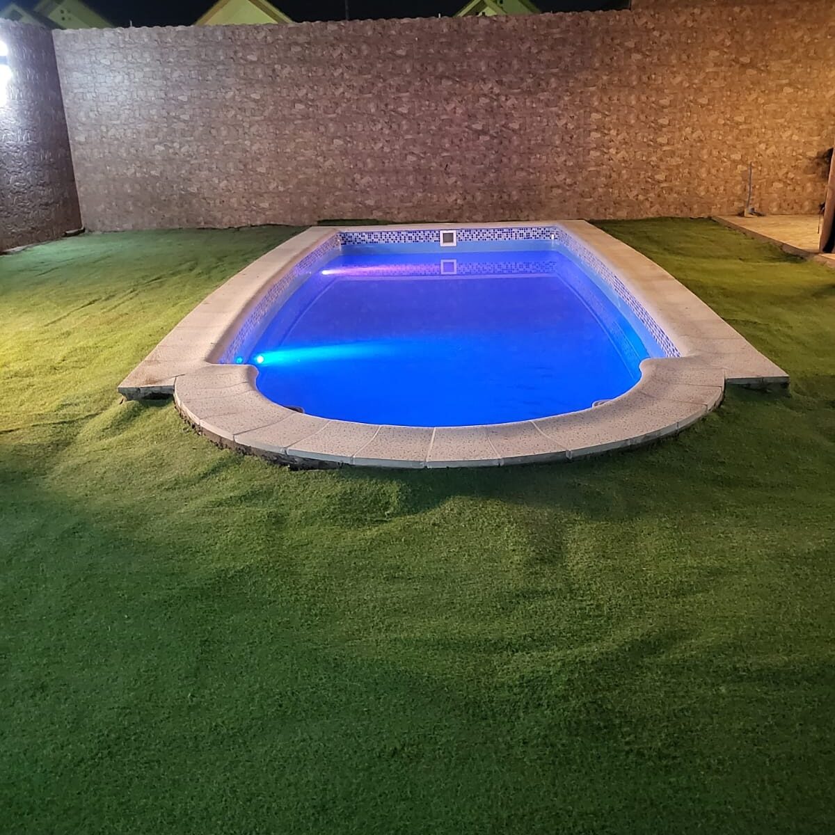 Piscina con iluminación LED azul instalada sobre pasto sintético, vista nocturna
